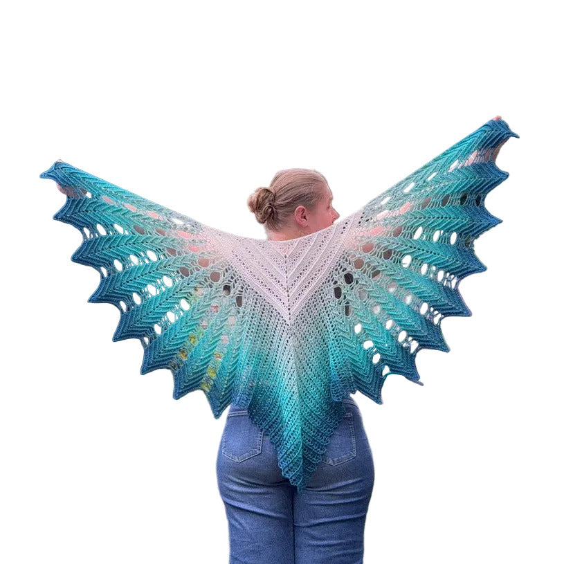 A woman viewed from behind wearing a large open-knit shawl with a gradient color story from white at the center to teal at the edges, paired with blue jeans against a plain white background.